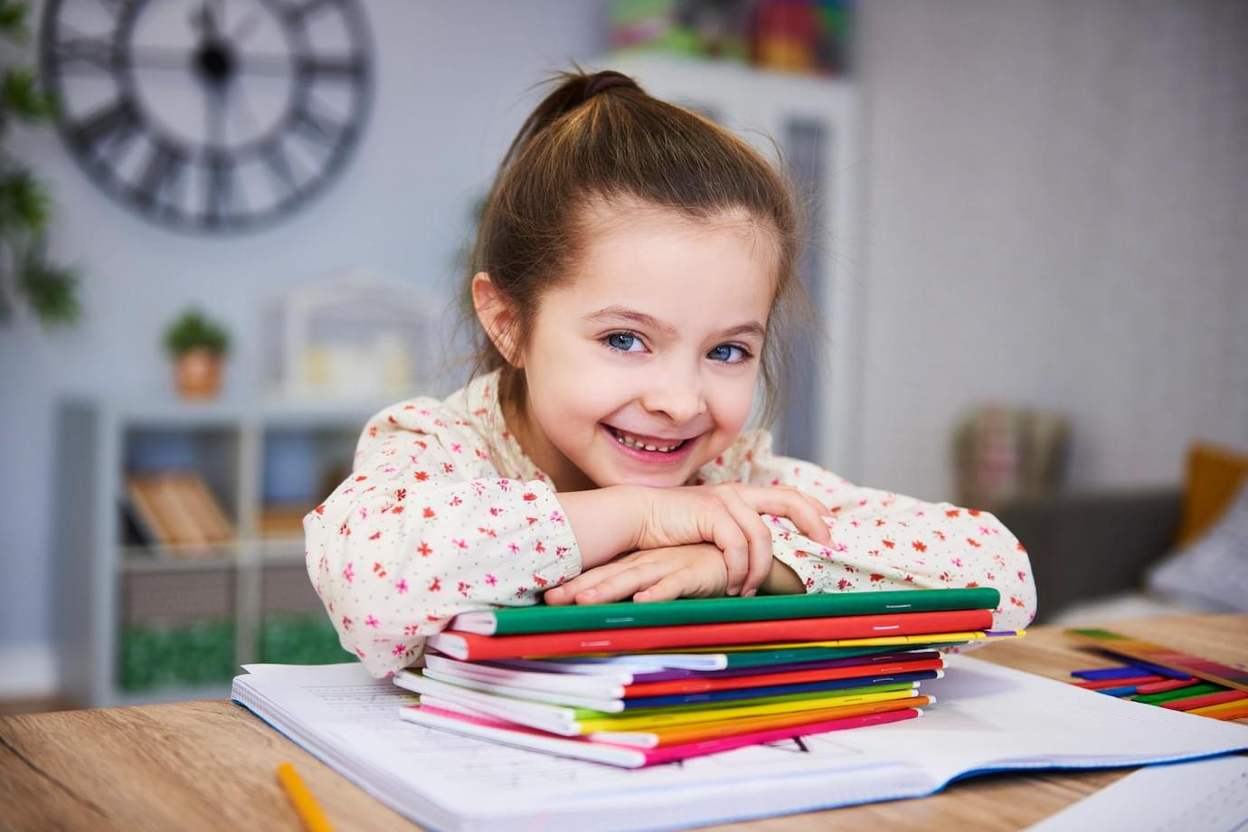 A child smiling over a stack of colourful notebooks at a desk