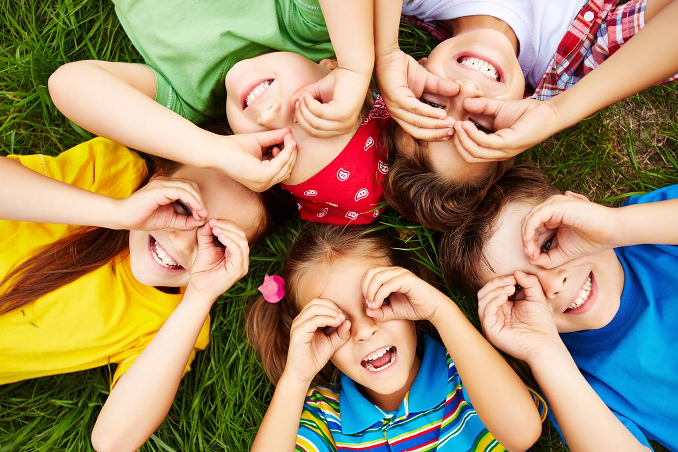 Children lying in a circle on the grass, looking through their hands like binoculars